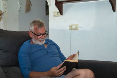 Olgun, sakallı, gözlüklü, pijamalı, evde kanepede oturmuş sessizce kitap okuyan beyaz saçlı bir adam. Kavram okuma, yalnızlık, emeklilik, emeklilik.