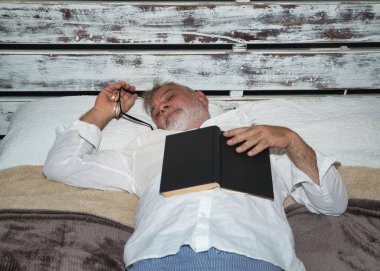 Olgun, gri saçlı, sakallı, elinde gözlüklü, pijamalı, üzerinde bir kitapla yatakta uyuyan bir adam. Kavram okuma, yalnızlık, emeklilik, emeklilik, uyku, uyku