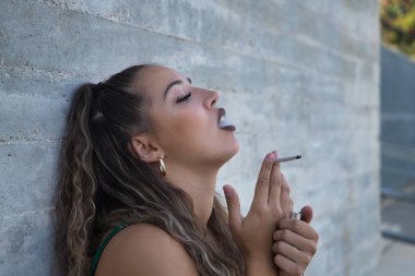 Latin ve İspanyol kız, genç ve uyumsuz, isyankar, tek başına sigara içiyor, dumanı şehvetli bir şekilde soluyor, gri bir arka planda. Kavram tütün, duman, ateş, bağımlılık