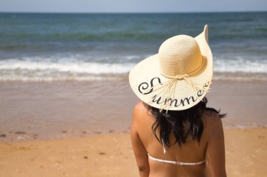 South American woman, young and beautiful, brunette with hat with the word summer and bikini on her back looking at the sea. Concept sea, sand, sun, beach, vacation, travel, summer.