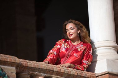 Tipik Fas kırmızısı takım elbiseli, altın ve gümüş ipliklerle işlenmiş İspanyol meydanındaki tuğla korkuluklara yaslanmış güzel bir genç kadın. Güzellik kavramı, etnik köken, tipik takım elbiseler, Arap.