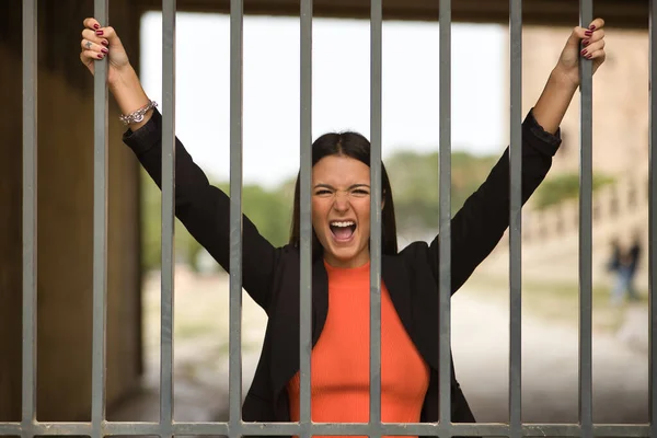Young beautiful woman with straight brown hair and jacket clinging to bars screaming furiously. Concept rebellion, screaming, problems, anxiety, millennial.
