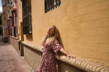 Orta yaşlı sarışın bir kadın, çiçekli bir elbise giyiyor, gözleri kapalı ve rahat güneşlenen bir duvara yaslanıyor. Kavram rahatlama, huzur, sükunet, mutluluk.