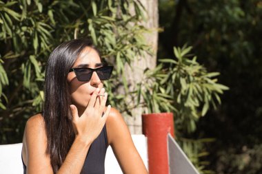 Genç bir kadın, esmer, ince, siyah giyinmiş, güneş gözlüğü takıyor, bankta oturmuş sigara içiyor. Kavram tütün, sigara, bağımlılık, sağlık, sigara.