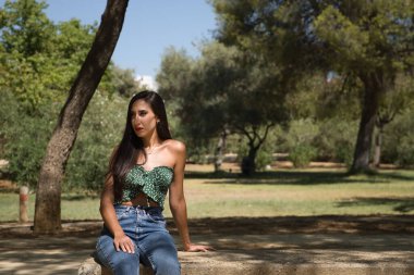 Genç bir kadın, esmer, ince, yeşil tişört ve kot giymiş, taş bir bankta oturuyor, yalnız, üzgün, ciddi. Yalnızlığı, kederi, üzüntüyü kavra..