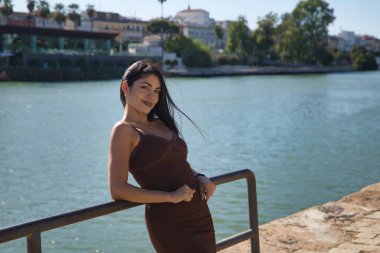 Güney Amerikalı bir kadın, genç ve güzel, esmer, kahverengi elbiseli, rahat ve çapkın bir tavırla korkuluklara yaslanmış. Moda, güzellik, trend, seyahat, gülümseme, rahatlama.