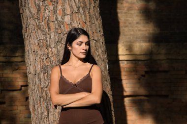 Güney Amerikalı bir kadın, genç ve güzel, esmer, kahverengi elbiseli, bir ağacın gövdesine yaslanmış, kollarını kavuşturmuş, kızgın. Öfkeyi, ciddiyeti, iğrenmeyi kavra..