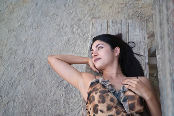 South American woman, young and beautiful, brunette, in leopard dress, lying on a wooden bench, relaxed and thoughtful. Concept beauty, fashion, tranquility, relax, think.