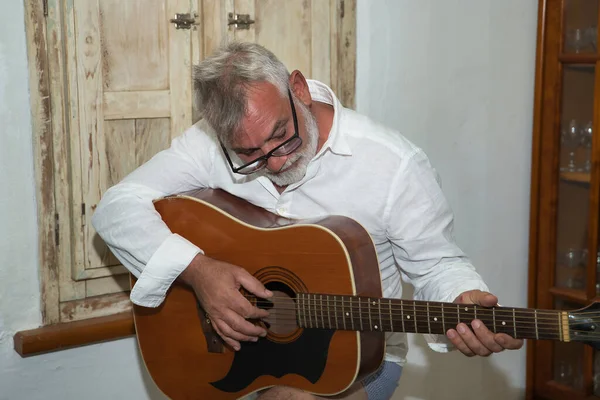 Mature man, gray-haired and bearded, with glasses, wearing a white shirt, happy to learn to play the guitar. Concept loneliness, retirement, pensioner, music, instruments, lessons.