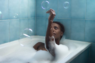 Köpük dolu bir küvetle banyo yapan ve sabun köpüğüyle oynayan Afro-Amerikalı bir kadın. Kavram banyosu, rahatla, köpük, sabun, baloncuk.