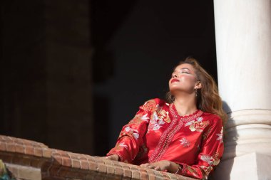 Tipik Fas kırmızısı takım elbiseli, altın ve gümüş ipliklerle işlenmiş İspanyol meydanındaki tuğla korkuluklara yaslanmış güzel bir genç kadın. Güzellik kavramı, etnik köken, tipik takım elbiseler, Arap.