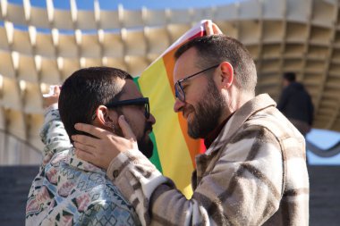 Eşcinsel bir çiftin gerçek evliliği, biri diğerinin yüzünü okşarken ve eşcinsel gurur bayrağı tutarken, mutlu ve suç ortağı. Kavram lgtb, lgtbiq +, çiftler, aşık, gururlu, bayrak.