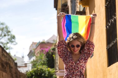 Middle-aged blonde woman, wearing a flowered dress and sunglasses, walking down the street and waving and a gay pride flag. Concept lgtbi, gay, lesbian, pride day. clipart