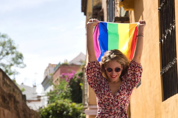 Orta yaşlı sarışın bir kadın, çiçekli bir elbise ve güneş gözlüğü takıyor, sokakta yürüyor, el sallıyor ve eşcinsel gurur bayrağı sallıyor. Kavram lgtbi, gay, lezbiyen, gurur günü.