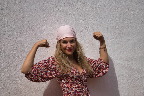 Middle-aged blonde woman, wearing a flower dress and a pink scarf of the fight against cancer, with arms up pulling muscle on a white background. Concept fight, disease, overcoming, cancer, breast.