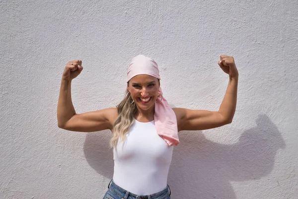 Young beautiful woman, with pink scarf of fight against cancer, with arms up pulling muscle, on white background. Concept fight, strength, disease, cancer, breast.