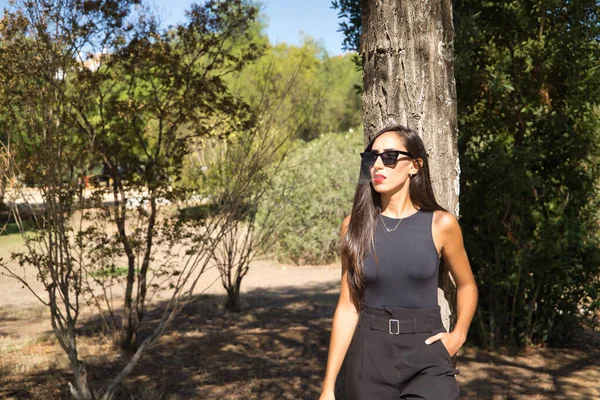 Genç bir kadın, esmer, ince, siyah giyinmiş ve güneş gözlüğü takıyor, sigara içerken bir ağaç gövdesine yaslanıyor. Kavram tütün, sigara, bağımlılık, sağlık, sigara.