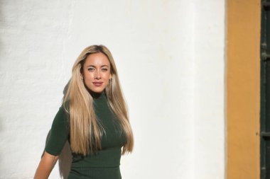 Young, beautiful, blonde woman with green eyes, wearing a green dress, leaning against a white wall, with seductive look. Concept beauty, fashion, trend, eyes, seduction.