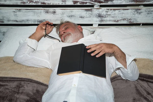 Mature, gray-haired, bearded man with a pair of glasses in his hand, in his pajamas, lying in bed asleep with a book on top of him. Concept reading, loneliness, retirement, pensioner, sleep, sleeping