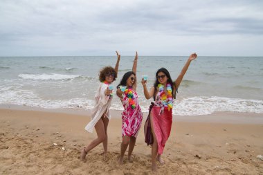 Attractive mature woman with two young, pretty, brunette South American women in bikinis, sunglasses and flower necklaces dancing with glasses of blue wine. Concept vacation, friends, summer, drinks.