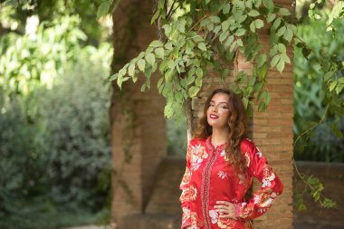 Altın ve gümüş ipliklerle işlenmiş tipik bir Fas kırmızı elbisesi içindeki genç ve güzel bir kadın, sarmaşıklı tuğla bir sütunun yanında poz veriyor. Güzellik kavramı, etnik köken, tipik takım elbiseler, Marakeş, Arap.