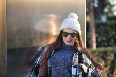 Young woman, beautiful, straight brown hair, sweater, coat, wool hat and sunglasses, leaning on a metal wall, playing with her hair, flirtatious and sensual. Concept beauty, autumn, winter, cold.