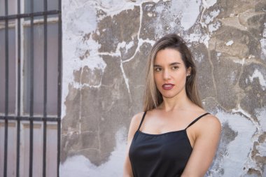 Young woman, beautiful and blonde, with black tank top, leaning against a wall with fallen paint, with a sensual and flirtatious look. Concept sensuality, beauty, flirting.