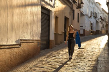 Young woman, beautiful and blonde, wearing a black tank top, blue fur jacket in her hand and jeans, walking down a lonely and lost street during the golden hour. Concept fashion, beauty, sunset.