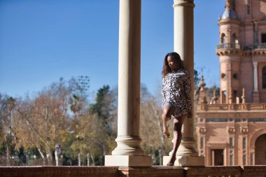 Young, beautiful, Latin and South American woman in short dress with zebra pattern, climbing a brick wall between two marble columns, posing sensual and attractive as a super model.
