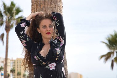 Genç ve güzel bir kadın, sarışın, kıvırcık saçlı ve mavi gözlü, çiçekli elbiseli ve siyah bluzlu, saçlarında elleri olan, şehvetli ve kışkırtıcı, palmiye ağacına yaslanmış..