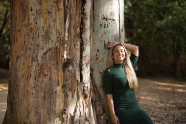 Young, beautiful, blonde woman with green eyes, wearing a green dress, leaning on the trunk of a big tree, relaxed, smiling and happy. Concept beauty, fashion, nature, autumn, relax.