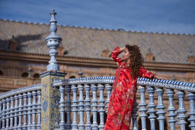 Altın ve gümüş ipliklerle işlenmiş tipik bir Fas kırmızı elbisesi içinde İspanyol meydanındaki fayanslı korkuluklara yaslanmış güzel bir genç kadın. Güzellik kavramı, etnik köken, tipik takım elbiseler, Arap.