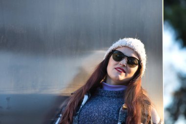 Güzel, düz kahverengi saçlı, kazak, ceket, yün şapka ve güneş gözlüklü, metal bir duvara yaslanmış, cilveli ve şehvetli bir kadın. Güzellik, moda, sonbahar, kış, soğuk kavramları.