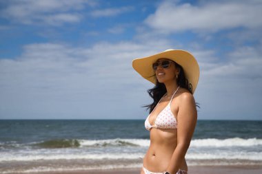 South American woman, young and beautiful, brunette with sunglasses, hat and bikini posing happy and smiling on the beach. Concept sea, sand, sun, beach, vacation, travel, summer.