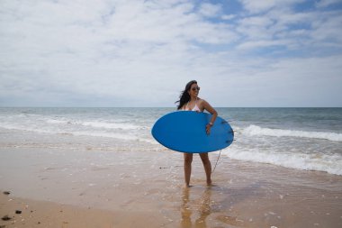 South American woman, young and beautiful, brunette with sunglasses and bikini, coming out of the water holding a blue surfboard. Concept sea, sand, sun, beach, vacation, surf, summer.