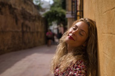 Orta yaşlı sarışın bir kadın, çiçekli bir elbise giyiyor, gözleri kapalı ve rahat güneşlenen bir duvara yaslanıyor. Kavram rahatlama, huzur, sükunet, mutluluk.