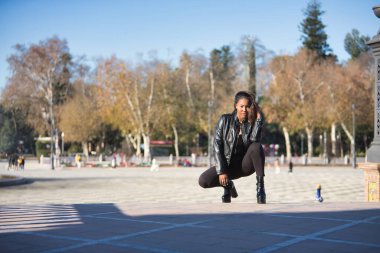 Young, beautiful, Latin and South American woman with leather jacket and top, jeans and platform shoes, squatting, posing sensual and attractive. Concept beauty, fashion, diversity, latina.