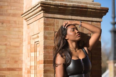Young, beautiful, Latin and South American woman with leather top and jeans leaning against a brick wall, touching her hair in a sensual and provocative attitude. Concept beauty, fashion, diversity.