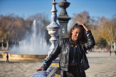 Genç, güzel, Latin ve Güney Amerikalı kadın deri ceketli, üst ve siyah kot pantolonlu fayanslı korkuluklara yaslanmış saçlarına baştan çıkarıcı ve flörtöz bir şekilde dokunuyor. Güzellik kavramı, çeşitlilik.