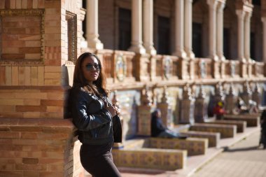 Young, beautiful, Latin and South American woman with leather top and jacket, sunglasses and jeans, leaning against a brick wall, posing sensual and attractive. Concept beauty, fashion, diversity.