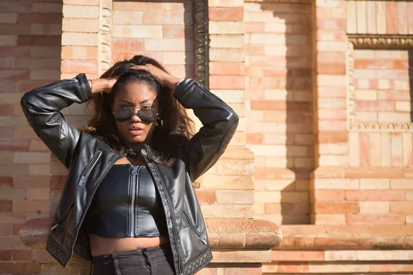 Portrait of young, beautiful, Latin and South American woman with leather top and jacket and sunglasses, leaning against a brick wall, hands in her hair, posing sensual and attractive.