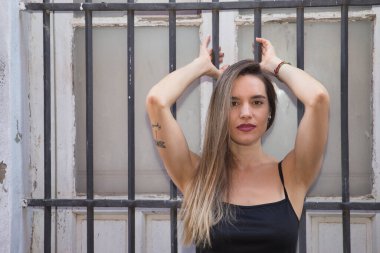 Genç bir kadın, güzel ve sarışın, siyah kolsuz tişörtlü, pencerenin ızgarasına yapışmış, şehvetli ve çapkın bakışlı. Duygusallık, güzellik, flört..