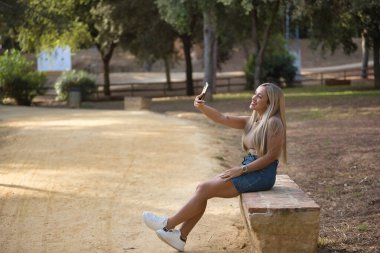 Genç, güzel, yeşil gözlü, tişörtlü, kot etekli, taş bir bankta oturan, cep telefonuyla fotoğraf çeken sarışın bir kadın. Cep telefonu kavramı, akıllı telefon, uygulamalar, sosyal ağlar.