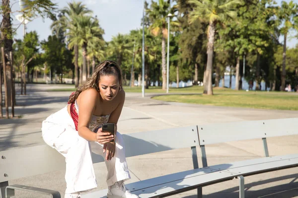 ラテン系とヒスパニック系の少女 若く美しい 彼女の携帯電話上のソーシャルネットワークをチェック ベンチに座って モバイルコンセプト スマートフォン コンサルティング アプリ — ストック写真