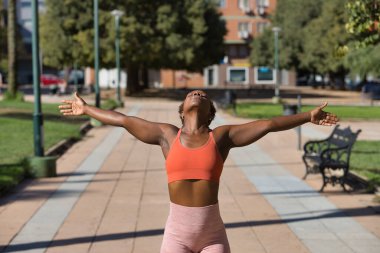 Spor giyimli Afro-Amerikan kadın kolları açık ve gözleri kapalı, rahatlamış ve hayata minnettar. Spor, meditasyon, sağlık, egzersiz, rahatlama.