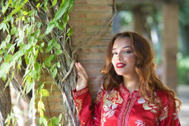 Altın ve gümüş ipliklerle işlenmiş tipik bir Fas kırmızı elbisesi içindeki genç ve güzel bir kadın, sarmaşıklı tuğla bir sütunun yanında poz veriyor. Güzellik kavramı, etnik köken, tipik takım elbiseler, Marakeş, Arap.