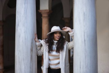 Young, beautiful, brunette woman with curly hair, beige sweater, coat and hat, leaning on marble columns, smiling and happy. Concept beauty, fashion, autumn, winter, cold, relax.