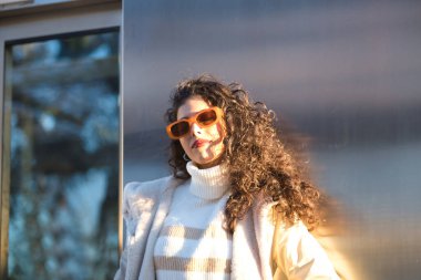 Young woman, beautiful, brunette with curly hair, sweater, coat and sunglasses, leaning on a metal wall, relaxed and calm receiving the sun's rays. Concept beauty, fashion, autumn, winter, cold, relax