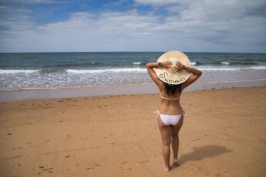 South American woman, young and beautiful, brunette with hat with the word summer held with both hands and bikini on her back looking at the sea. Concept sea, sand, sun, beach, vacation, travel.