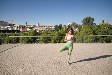 Orta yaşlı sarışın kadın, yeşil tayt ve beyaz bluz giyiyor, meditasyon yapıyor ve dışarıda yoga egzersizleri yapıyor. Yoga, meditasyon, konsantrasyon, spor, farkındalık.